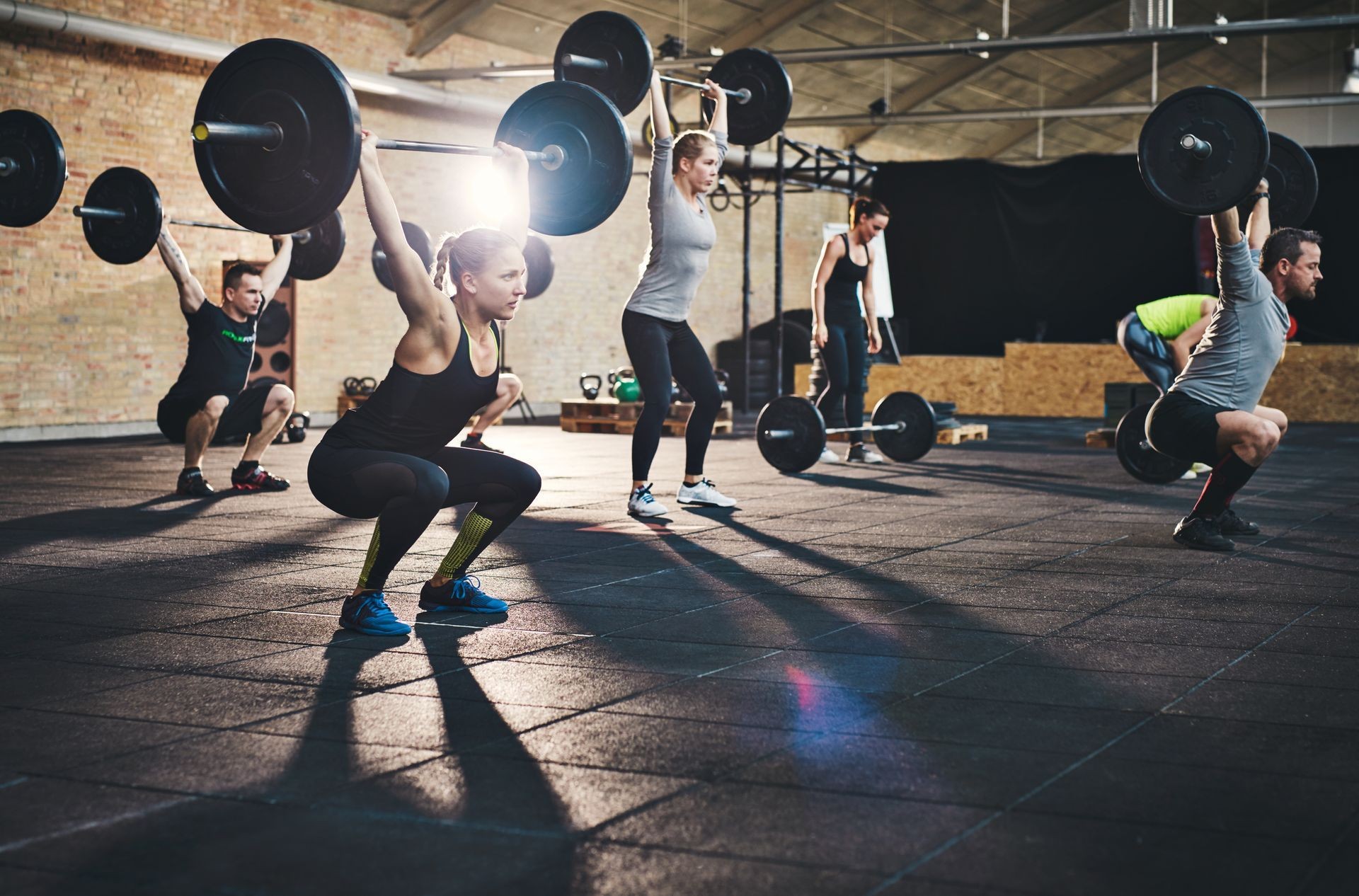 Group of people performing barbell overhead squats in a gym with high ceilings and exercise equipment.